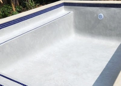 Finished and still empty small concrete swimming pool, with white interior, some blue tiles and a light.