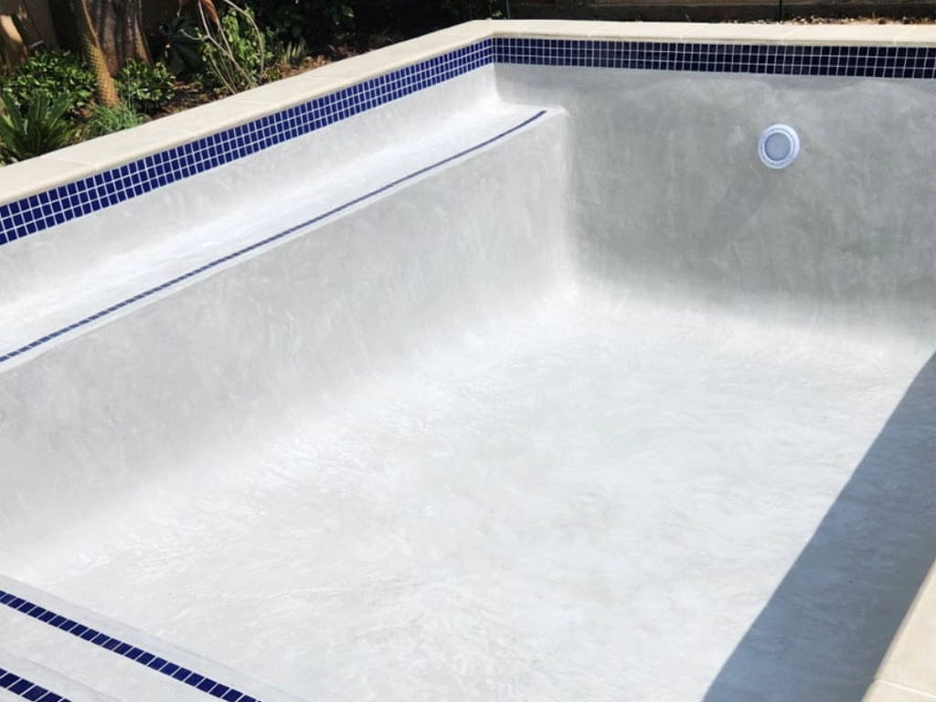 Finished and still empty small concrete swimming pool, with white interior, some blue tiles and a light.